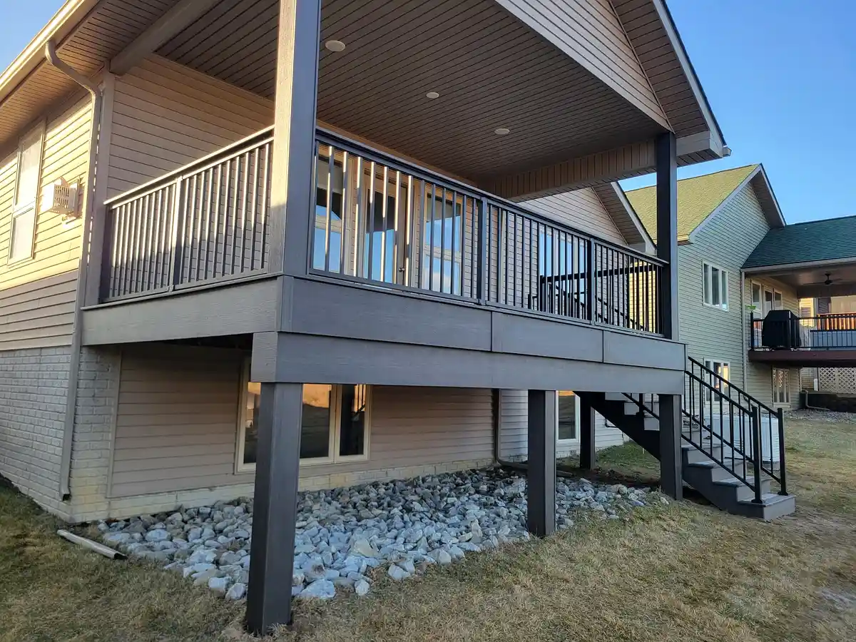 A large elevated composite deck with charcoal gray decking, black metal railings, and a covered porch area with recessed ceiling lights and open soffits. A side staircase with matching railings provides yard access — an excellent example of a high deck remodel for Philadelphia homes.