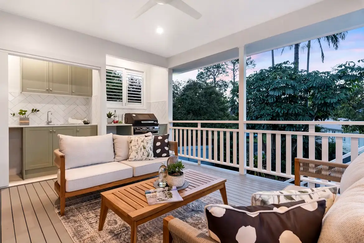 A stunning covered outdoor deck featuring a built-in outdoor kitchen with sage green cabinetry, herringbone tile backsplash, and a gas grill, paired with a cozy lounge seating area and ceiling fan. Open railings frame a lush garden view at dusk, creating the perfect outdoor living space in Philadelphia.