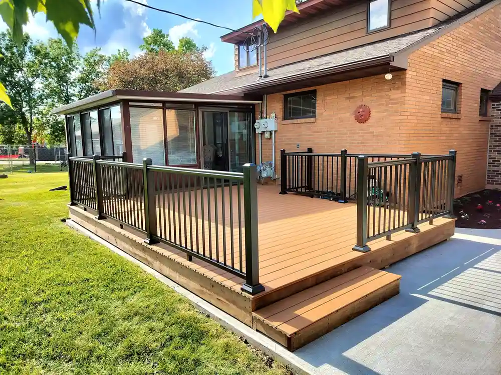 A freshly built ground-level composite deck with warm cedar-tone boards and sleek black aluminum vertical railings, connected to an enclosed sunroom addition on a brick home. Steps lead down to a concrete patio, making this a perfect outdoor entertainment space for Philadelphia homeowners looking for a deck builder.