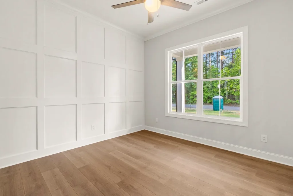 A freshly built home addition room featuring elegant white box-panel wainscoting accent wall, warm oak hardwood floors, crown molding, and a large window with views of lush green trees. A clean, timeless interior finish that adds premium square footage to any Philadelphia home addition project.