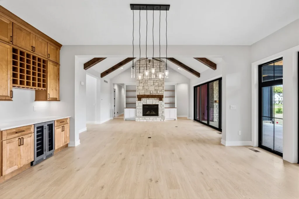 A spacious open-concept home addition living area featuring vaulted ceilings with dark wood beams, a natural stone fireplace, a built-in bar with wine fridge and wine rack cabinetry, and floor-to-ceiling black sliding doors. A high-end Philadelphia home addition built for modern family living and entertaining.