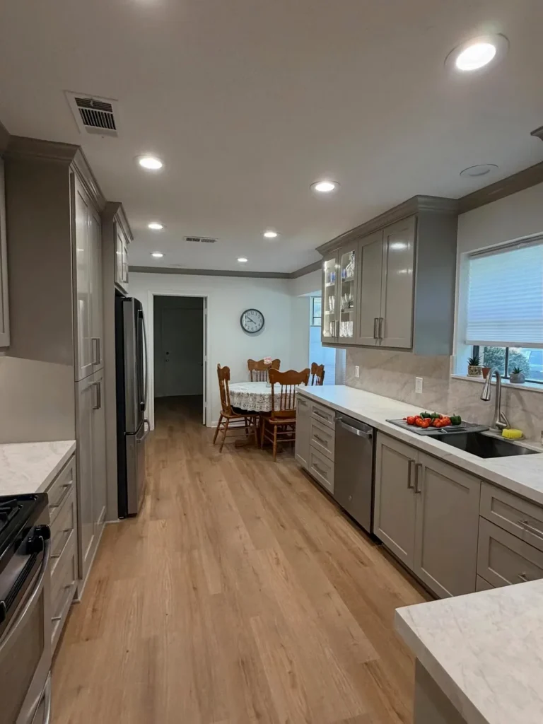 A beautifully remodeled galley-style kitchen featuring grey shaker cabinets, white quartz countertops, and a marble-look tile backsplash with stainless steel appliances. The warm hardwood floors and recessed lighting create a bright, open feel that flows into the dining area. A stunning kitchen remodel result for Philadelphia homeowners looking for a modern yet cozy upgrade.